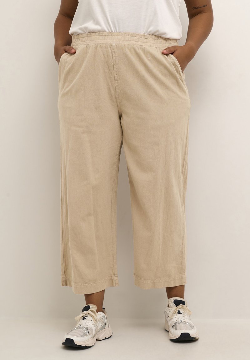 Personne portant un pantalon beige ample raccourci avec taille élastique et des baskets blanches, debout les mains dans les poches.