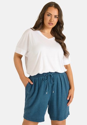 Kvinde med langt brunt hår iført en løs hvid V-hals T-shirt og blå shorts med snor, stående med hænderne i lommerne.