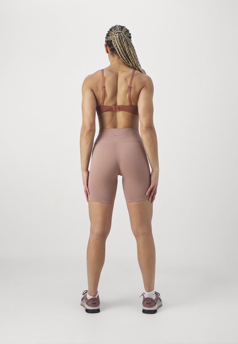 Femme aux cheveux tressés, debout dos à la caméra, portant un soutien-gorge de sport, un short taille haute et des baskets dans des tons roses doux sur un fond uni.
