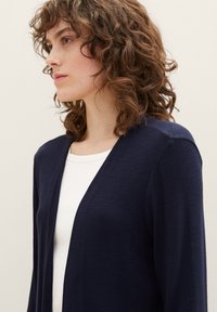 Marineblauwe cardigan van een lichtgewicht breiwerk, met een open voorkant, lange mouwen en een relaxte pasvorm, gecombineerd met een wit topje eronder.