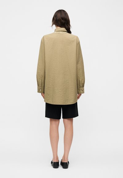 Άτομο με μακριά καστανά μαλλιά φοράει μια υπερμέγεθη μπεζ υφασμάτινη μπλούζα, μαύρα σορτς μέχρι το γόνατο και μαύρα επίπεδα παπούτσια, με την πλάτη γυρισμένη.