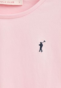 Camiseta de algodón rosa claro con cuello redondo; presenta una pequeña figura bordada en azul marino sosteniendo un bastón en el lado izquierdo del pecho.