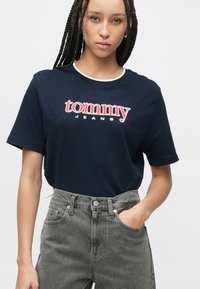 Tmavomodré bavlnené tričko s logom "tommy Jeans" v červenej a bielej farbe na hrudi. Kombinované s sivými riflemi s vysokým pásom.