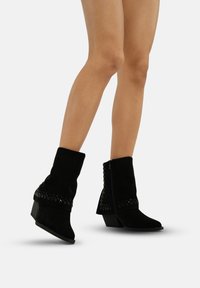Botines de ante negros con tacón cuadrado, que presentan detalles trenzados y acentos texturizados a los lados, exhibidos en las piernas de un modelo.