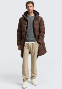 Brauner gefütterter Mantel mit Kapuze, Reißverschluss und seitlichen Taschen. getragen über einem grauen Pullover und beigen Hosen, mit weißen Turnschuhen.