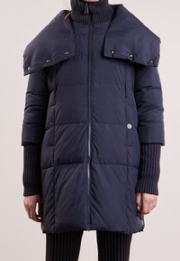 Kvinna klädd i en lång navyblå vadderad kappa med hög krage och tryckknappslucka över axlarna, tillsammans med svarta ribbade ärmar och randiga byxor.