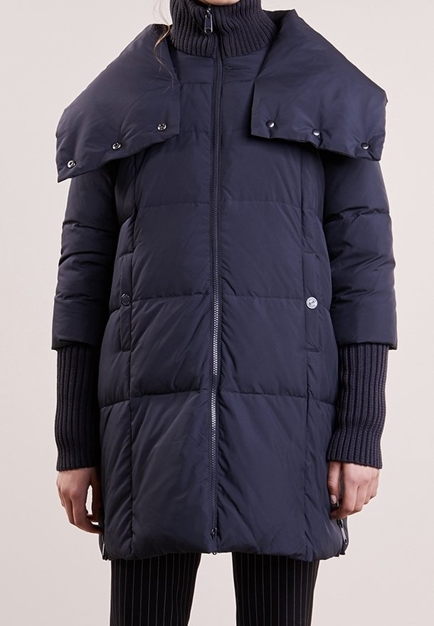 Kvinna klädd i en lång navyblå vadderad kappa med hög krage och tryckknappslucka över axlarna, tillsammans med svarta ribbade ärmar och randiga byxor.