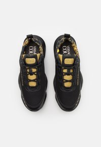 Chaussures de sport noires avec des accents jaunes, dotées d'un panneau latéral texturé, de lacets noirs et d'une doublure intérieure à motifs. Branding "VERSACE COUTURE" visible.