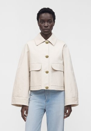 Persona de piel oscura que lleva una chaqueta abotonada color crema con grandes bolsillos y mangas anchas, combinada con vaqueros azul claro.