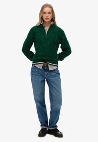 Groene gebreide trui met een ritssluiting bij de kraag, gecombineerd met een blauw-wit gestreept shirt, losse blauwe jeans en donkere schoenen.