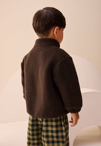 Veste en polaire marron à col montant, associée à un pantalon à carreaux verts et bleus, vue de dos. Texture douce, coupe décontractée.