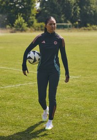 Čierny a tyrkysový športový komplet s oranžovými pruhmi, drží čierno-biely futbalový loptu a stojí na zelenej tráve.