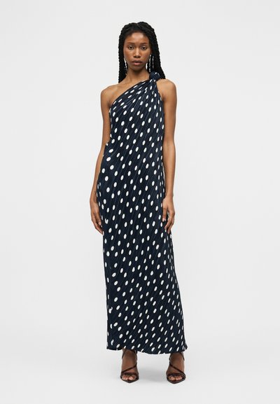 Donna con lunghe trecce che indossa un vestito maxi blu navy senza maniche con pois bianchi e sandali neri con cinturini, in piedi davanti a uno sfondo neutro.