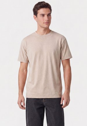 Junger Mann, der ein beiges kurzärmeliges T-Shirt und dunkle Jeans trägt, vor einem schlichten weißen Hintergrund steht und leicht zur Seite schaut.