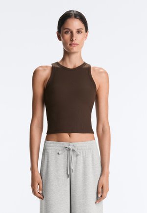 Kvinde med mørkt hår iført en brun ærmeløs crop top og lysegrå bukser med snøre, stående mod en ensfarvet hvid baggrund.