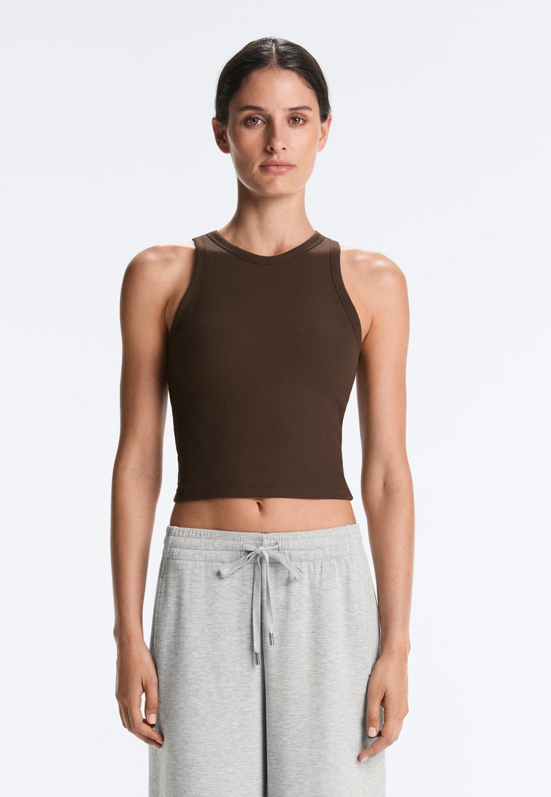 Femme aux cheveux foncés, en crop top marron sans manches et pantalon gris clair à cordon, debout devant un fond blanc uni.