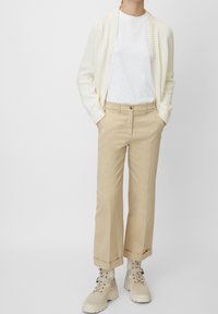 Persona che indossa un cardigan lavorato a maglia color crema, una t-shirt bianca, pantaloni beige con risvolti e stivali alti beige con lacci, in piedi con le mani in tasca.