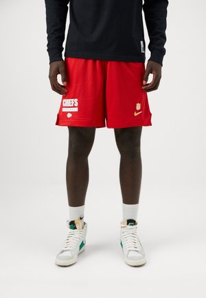 Man draagt rode sportshorts van Kansas City Chiefs, witte Nike high-top sneakers en een zwart shirt met lange mouwen, staand op een witte vloer.