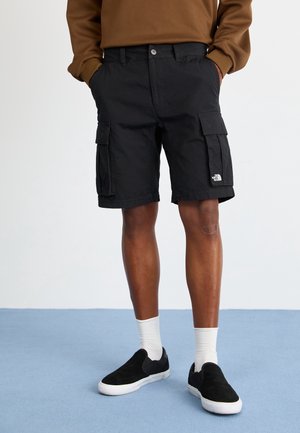 Man draagt zwarte cargoshorts, witte sokken en zwarte slip-on sneakers, staat met handen in de zakken op een blauwe vloer.