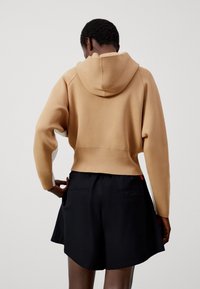 Przycięta bluza z kapturem z beżowej tkaniny z długimi rękawami i białymi akcentami, w zestawie z czarnymi szortami z tylnymi kieszeniami.