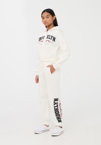 Biała kurtka z kapturem na zamek błyskawiczny i pasujące dresy. Z przodu i z boku znajdują się czarne i czerwone napisy "BROOKLYN". Gładki materiał, luźny krój.