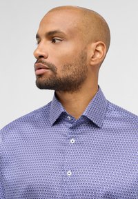 Blauw patroon overhemd met een gladde textuur, voorzien van een kraag en knoopsluiting. Het ontwerp bevat een herhalend druppelpatroon in een donkerder blauw.