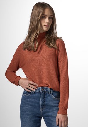 Langsleeve high-neck trui in roestkleur, met een gestructureerde stof, gecombineerd met blauwe spijkerbroek, heeft een losse pasvorm en opgerolde mouwen.