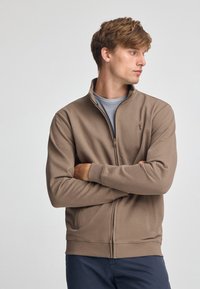 Felpa zip-up marrone con colletto, maniche lunghe e tasche laterali; presenta un piccolo logo sul petto sinistro. Tessuto dalla consistenza liscia.