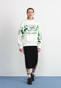 White sweatshirt with green checkered design, "THE ACES" logo, black midi skirt, and black sneakers with red accents and white socks.