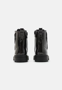 Botas de charol negro con un acabado brillante, dos tiradores y una suela de goma negra gruesa. La vista trasera muestra detalles de textura y diseño.