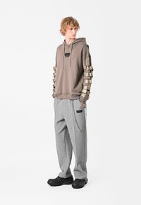 Sudadera marrón con un diseño texturizado, que presenta acentos en beige en las mangas. Pantalones de chándal grises con una cadena lateral y un parche con logo. Calzado negro.
