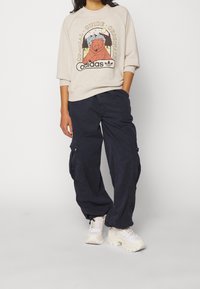 Beige sweatshirt med djurtryck och text, kombinerat med marinblå cargobyxor med sidofickor och vita klumpiga sneakers.