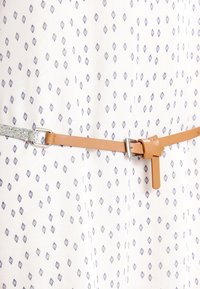 Hnědý kožený opasek se stříbrným kováním, stahující vzorovaný oděv s modrými diamantovými tvary na bílém pozadí. Hladký povrch.