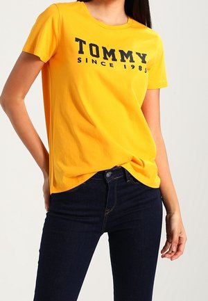 Person iført en skarp gul kortærmet T-shirt med teksten "TOMMY Since 1985", kombineret med mørkeblå jeans mod en ensfarvet baggrund.