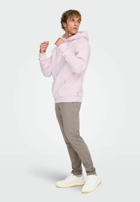 Felpa con cappuccio rosa chiaro in tessuto morbido, con tasca frontale e cappuccio con coulisse regolabile. Abbinata a pantaloni jogger affusolati grigio chiaro e sneakers bianche.