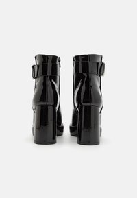 Botas de tobillo de charol negro con puntera redondeada, correa ajustable con hebilla y tacón grueso. Textura suave con un acabado brillante.