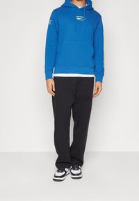 Modrá mikina s kapucí, předním kapsou a žlutým logem, kombinována s černými kalhotami. Obsahuje bílý tričko a černé s bílými teniskami.