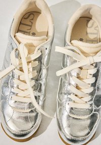 Pár stříbrných metalických tenisek s bílými šňůrkami a béžovým polstrovaným jazykem na světlém povrchu.