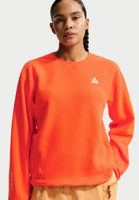 Γυναίκα με πλεξούδες φοράει μια φωτεινή πορτοκαλί μακρυμάνικη φλις μπλούζα με λευκό τρίγωνο λογότυπο στο στήθος και μπεζ παντελόνι.