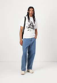 White graphic t-shirt featuring black angular print, paired with loose-fitting light blue denim jeans and beige sneakers. Black shoulder strap bag.