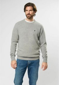 Hombre con barba que lleva un suéter de cuello redondo gris claro y jeans azules, de pie contra un fondo claro y liso.