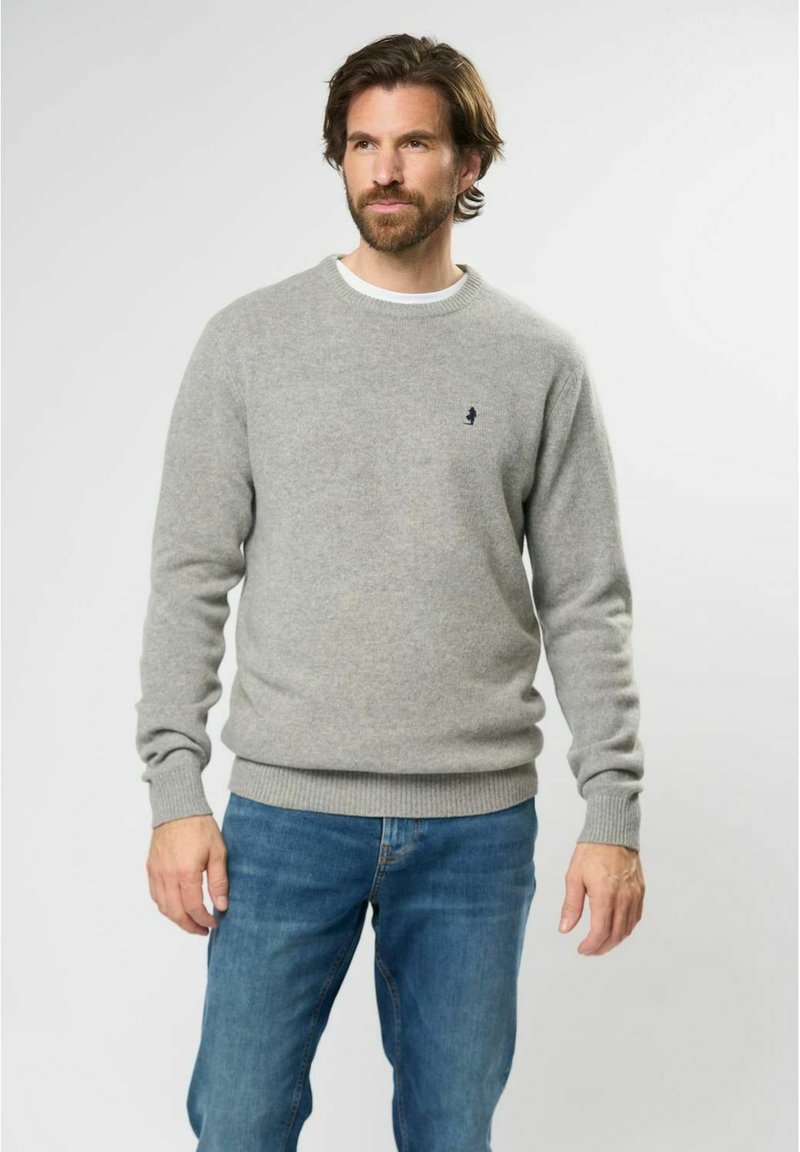 Hombre con barba que lleva un suéter de cuello redondo gris claro y jeans azules, de pie contra un fondo claro y liso.