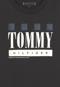 T-shirt nera in cotone, con grande scritta bianca "TOMMY HILFIGER" e due accenti quadrati blu sopra il nome del marchio.