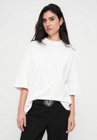 Λευκό υπερμεγέθες μπλουζάκι με στρογγυλή λαιμόκοψη, κοντά μανίκια και λεία υφή, συνδυασμένο με μαύρο παντελόνι και ασημένια καρδιά αγκράφα ζώνης.