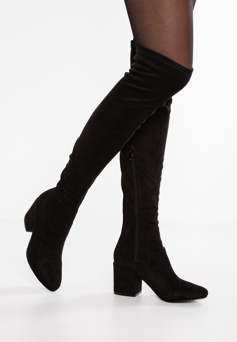 Botas negras de ante por encima de la rodilla con tacones en bloque y cremallera lateral, usadas con medias negras transparentes sobre fondo blanco.