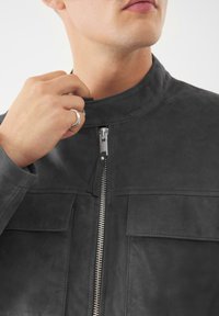Giacca in suede nero con colletto rialzato, zip frontale verticale e due tasche sul petto con patta. La mano del modello stringe il colletto.