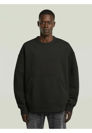 Giovane uomo che indossa una felpa nera oversize semplice con tasca frontale e jeans grigio scuro sbiaditi su sfondo grigio chiaro.