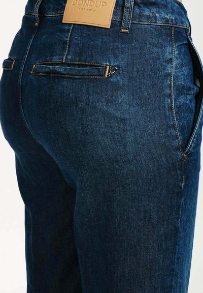 Jeans in denim blu scuro con un design aderente, una texture sottile e cuciture arancioni a contrasto. Presentano dettagli sulla tasca posteriore e un'etichetta in pelle.