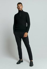 Gebreide zwarte trui met hoge kraag en knoopsluiting, gecombineerd met zwarte slim fit broek en zwarte schoenen, tegen een neutrale achtergrond.