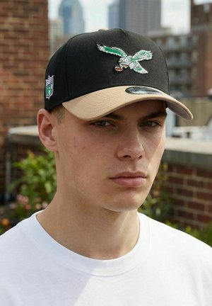 Junger Mann, der eine schwarze und beige Baseballkappe mit einem grünen Adler trägt, der einen Football hält, bestickt darauf, und der im Freien in der Nähe einer Backsteinmauer steht.